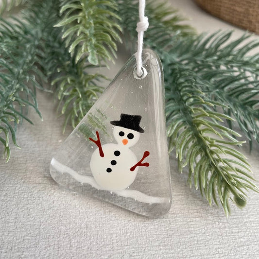 A triangular clear glass ornament featuring a painted snowman with a black top hat, orange nose, and red arms, hanging against green pine branches on a light fabric background.